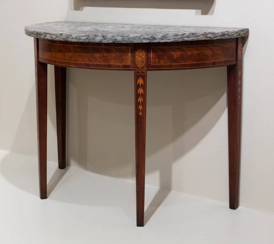 A half-moon console table with a grey marble top, an inlaid mahogany apron and tapered legs