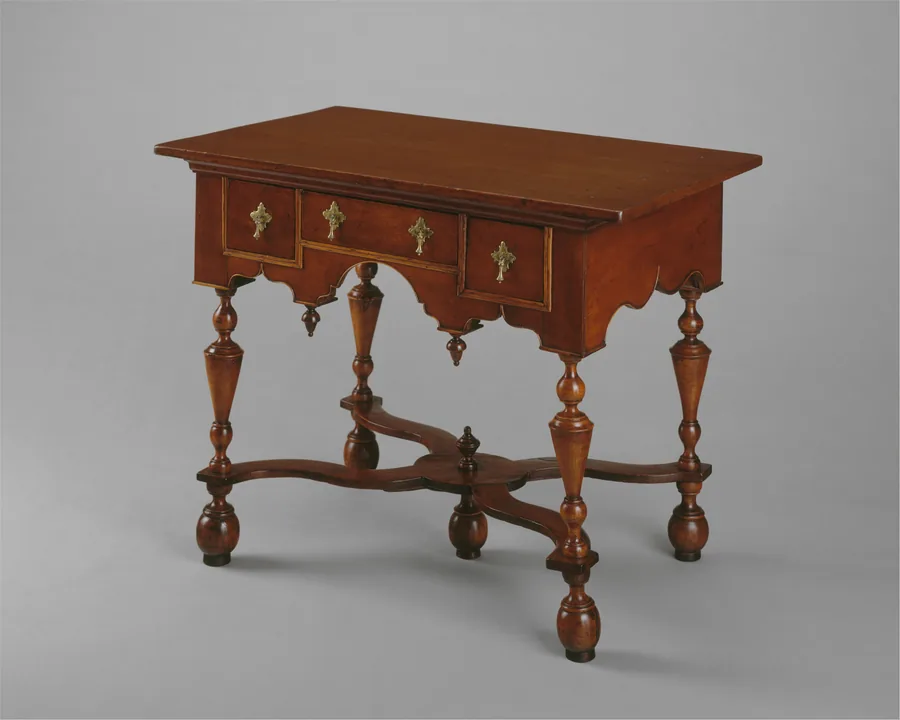 A walnut side table with three drawers, a scalloped apron with two drop finials, trumpet-turned legs and an X-shaped stretcher