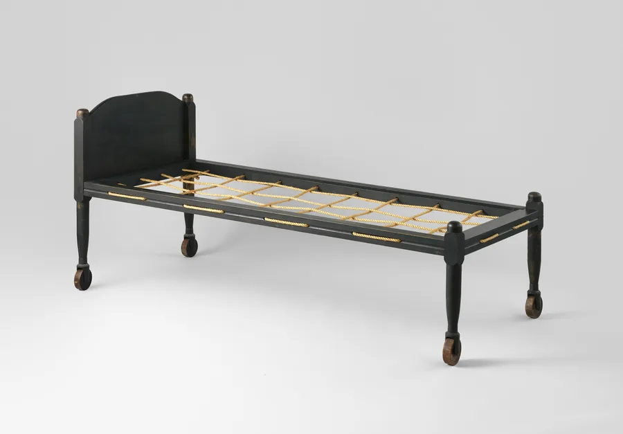 A plain dark-painted single bed frame with a low arched headboard, a rope deck and legs on castors