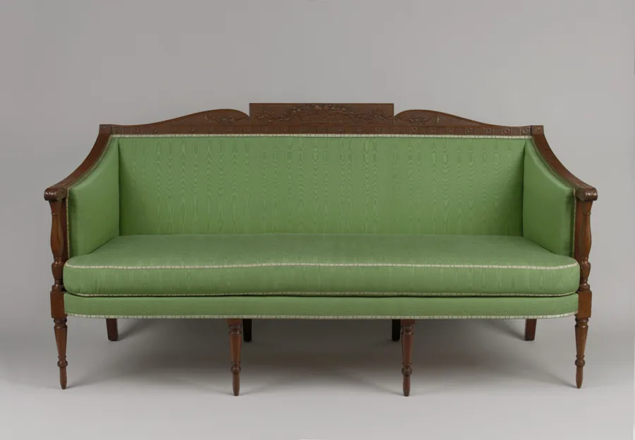 A Sheraton-style three-seat sofa with green upholstery, a carved crest rail and turned legs