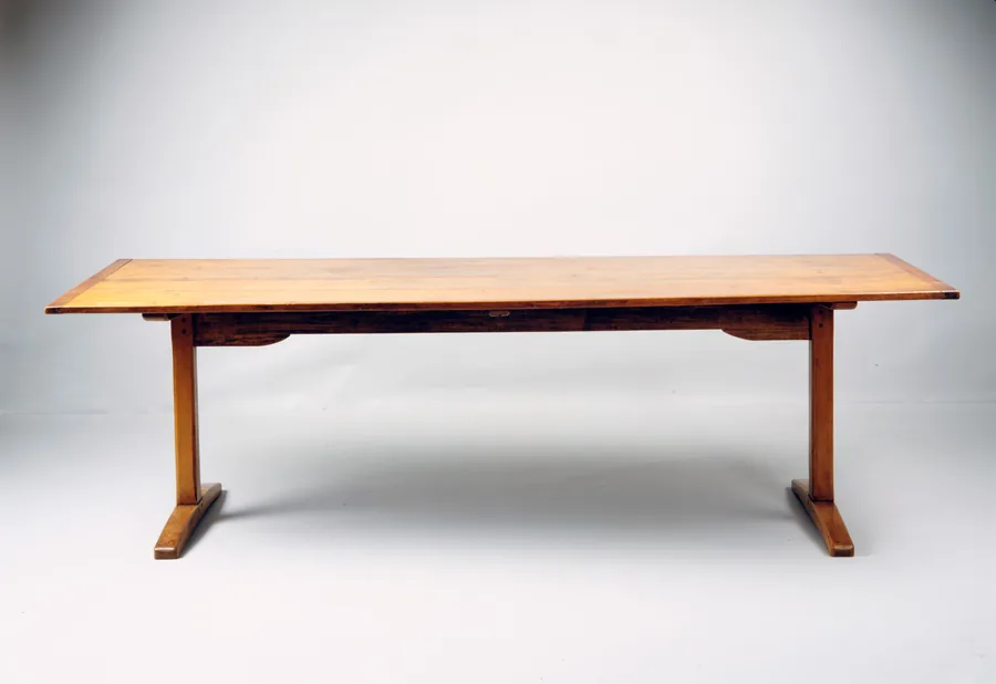 A long plain wooden dining table on two T-shaped trestle legs joined by a stretcher